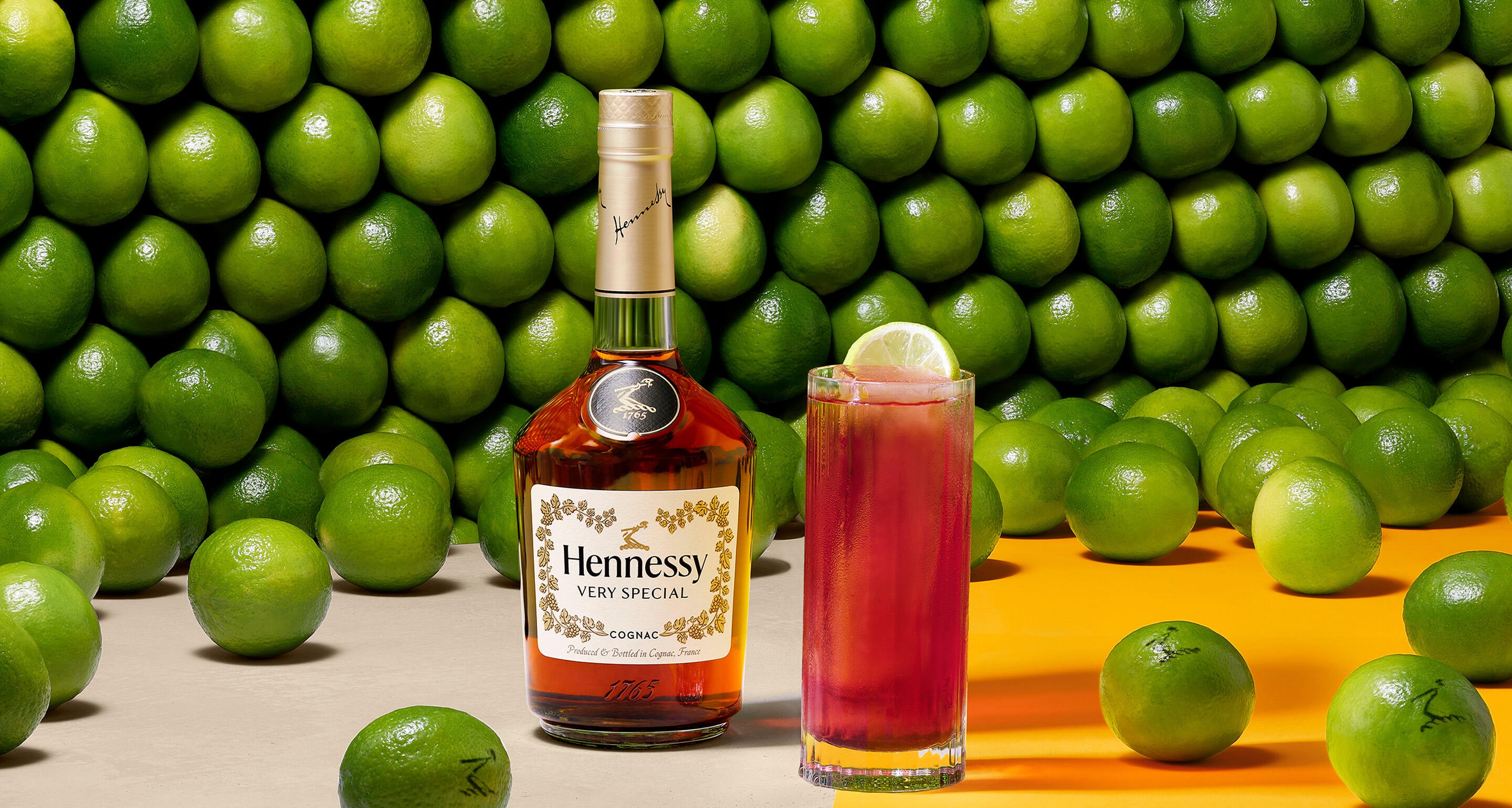 Bouteille de cognac Hennessy VS devant un mur de citrons verts, à côté d'un cocktail Hennessy Berry garni d'une tranche de citron vert