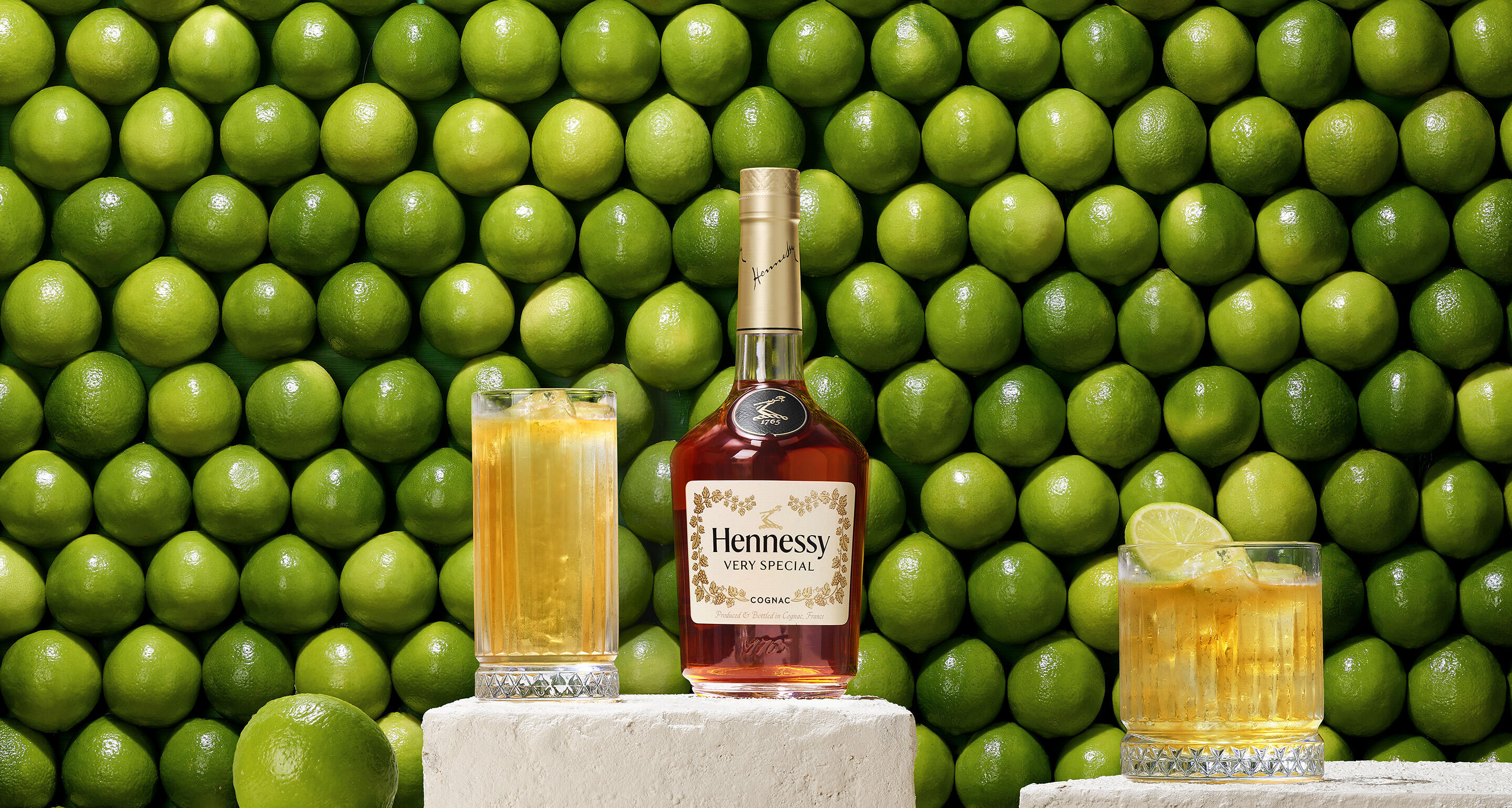 Bouteille de cognac Hennessy VS devant un mur de citrons verts, à côté de 2 cocktails Hennessy ginger servis avec une tranche de citron vert