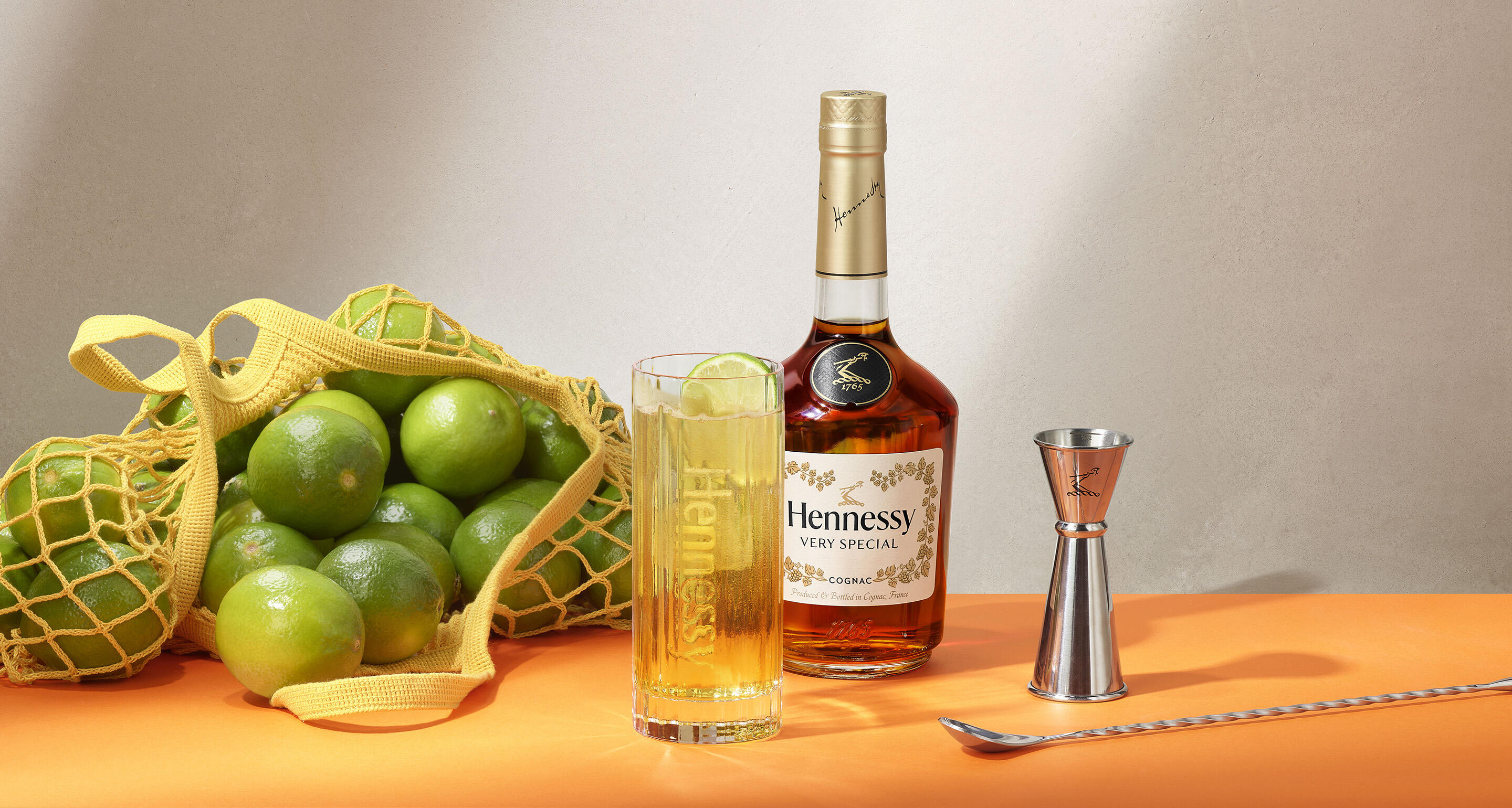 Bouteille de cognac Hennessy VS, à côté d'un filet de citrons verts frais et d'un cocktail servi dans un verre highball, avec des glaçons