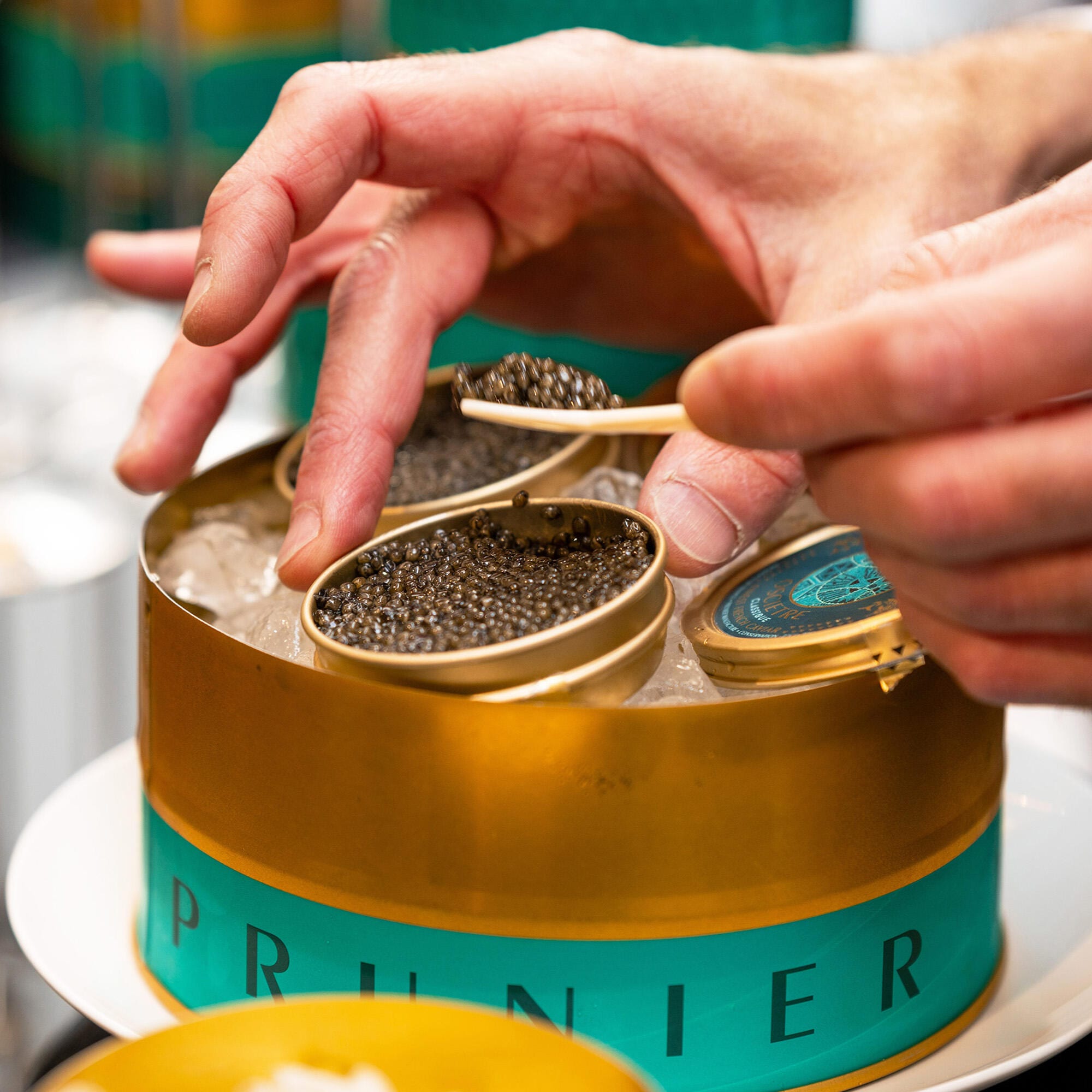Gros plan sur la main d'un client au-dessus d'une boite de caviar Prunier, tenant une petite cuillère en nacre blanche remplie de caviar