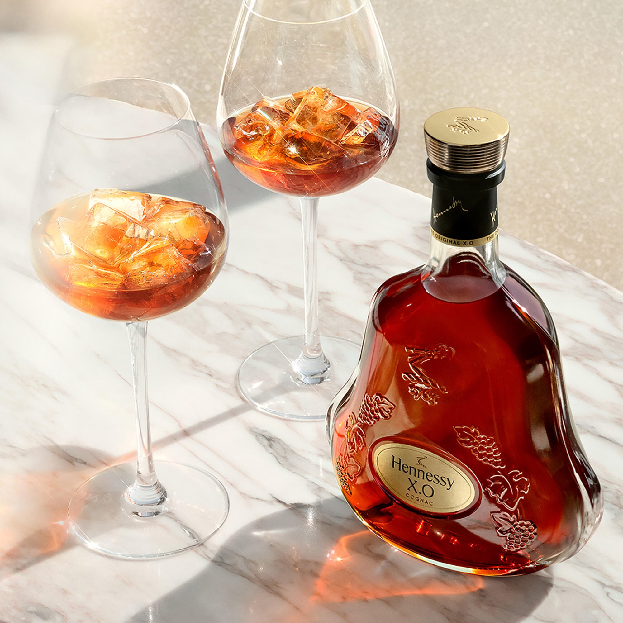 Carafe de cognac Hennessy X.O sur une table basse, à côté de 2 verres de cognac servis avec des glaçons
